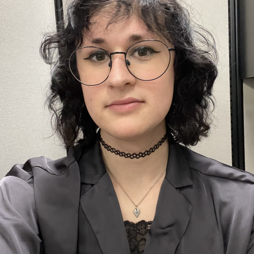 A portrait of Hayley, she is wearing a gray blazer with a lace top, silver pendant necklace and a black choker and has black wire-rimmed glasses.