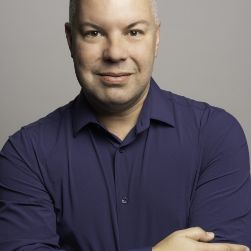 Jacob Brent in a blue shirt against a gray background with his arms folded over his chest