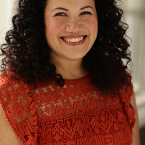 headshot of Jennica Carmona seen smiling at the viewer. She has long curly black hair and is wearing a crimson embroidered top with short sheer sleeves.