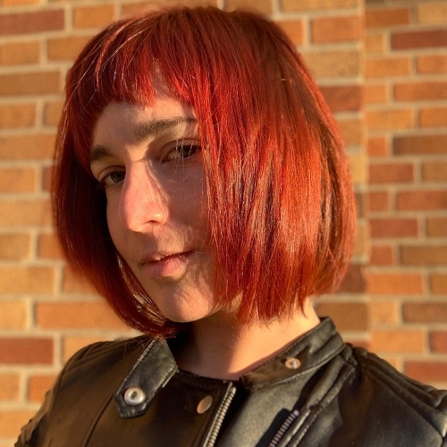 portrait of Katherine Freer wearing a black leather jacket, with red hair, against a brick background in sunset light
