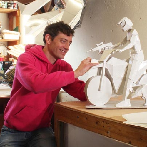 Nick Lenker works on a white ceramics piece of a subject on a bike holding a gun in his studio