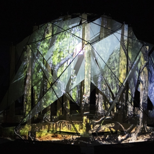 A tent featuring projections of the forest and abstracted structures on a dark stage