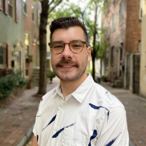 headshot of michael lambui. he is standing in a narrow tree-lined street of brick houses. Michael is wearing a white short-sleeved collared shirt with dark blue paint streaks. he has a short mustache and horn-rimmed glasses. 
