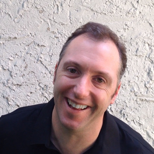Mike Worth headshot, smiling, black shirt, in front of white stucco wall