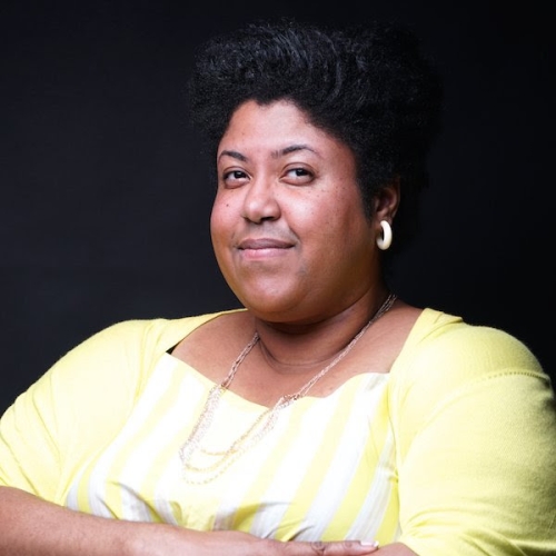 Monica O. Montgomery with her arms folded in a yellow and white shirt against a black backdrop