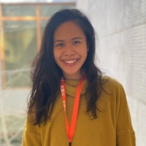 headshot of Nhi Nguyen. She is standing against a blurry background of a grey brick wall and is smiling at the camera. she has long dark hair and is wearing a chartreuse turtleneck sweater