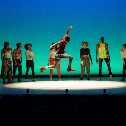 A group of dancers stand in a semi circle as a male dancer jumps in a spotlight in the center.