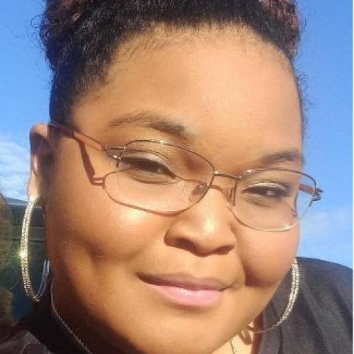 headshot of Nevaeh Ryals, with slim gold glasses, lark sparkling hoop earrings, and a glittering butterfly pendant on a small chain. Nevaeh is posed against a bright clear blue sky 