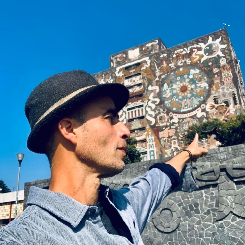 Rodrigo Esteva Ramirez looking away from the camera and wearing a blue shirt and dark gray fedora and pointing to a mural on a building up and behind him