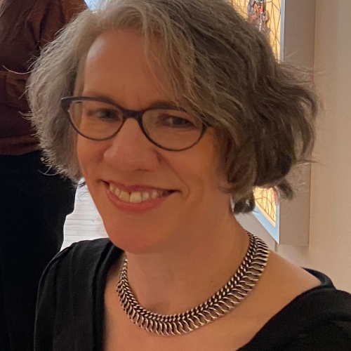 A closeup of Judith Schaechter wearing a silver necklace and a black shirt in what looks like a gallery space