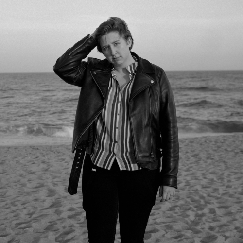 Keavy Handley-Byrne in a leather jacket and standing on a beach with their hand on their head 