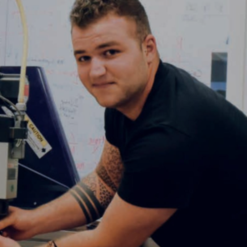 Shane Mastronardi, industrial design student, working with 3-D printer, black t shrt, tattoos on right arm