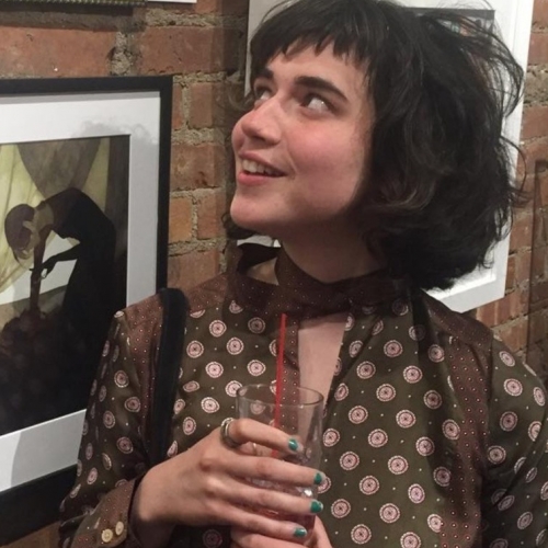 Illustration student Shannon Ryan in front of brick wall covered with framed illustrations, black hair, smiling, brown dress with white circles, holding glass with red straw