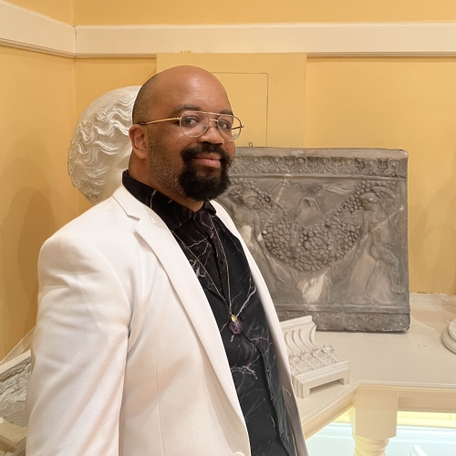 Darryl Babatunde Smith wearing a white blazer and a black shirt and standing in front of two pieces of ancient art in a room with yellow walls