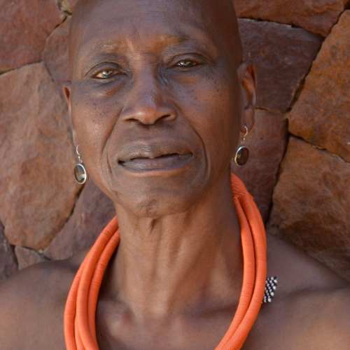 Germaine Acogny in an orange fiber necklace and standing in front of what looks like a deep brown rock structure