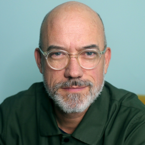 headshot of trey lyford against an pale blue background on a chartreuse couch. Trey is wearing a dark green buttondown shirt. Trey has a sly smirk under his grey trim beard. he is bald and is wearing clear acrylic glasses. 