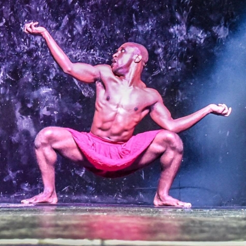 Uwazi Zamani shirtless in a pink skirt squatting with one arm raised and one arm resting on his knee