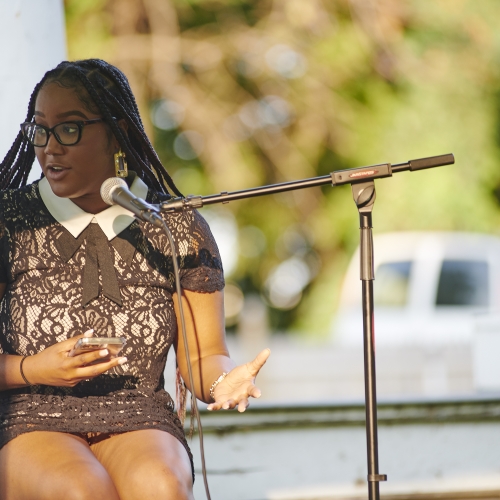 Zindzi Harley, UArts Museum Studies MA graduate, seated at an outdoor stage and speaking into a microphone. she is wearing black glasses and is looking to her right, and is wearing a brown lacey dress. 