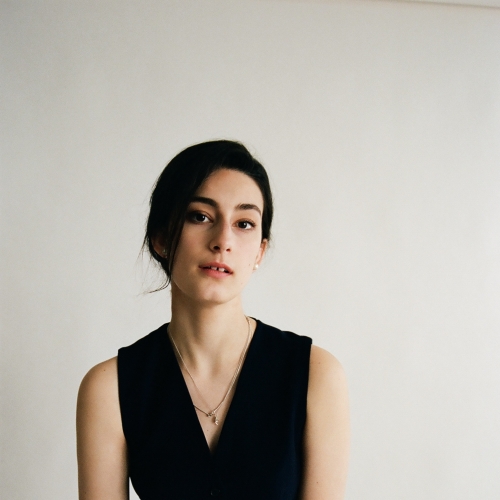 portrait of zoe nebraska feldman, pictured against a blank background and seated slightly on a stool. Zoe is wearing a black romper with a deep v neck with small pendants. zoe's hair is largely over the left side of her face and she has a serious expression with her mouth slightly open. 