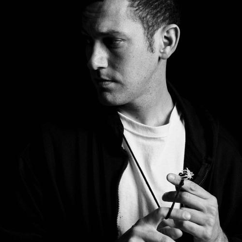 Alex Da Corte in black and white looks off to the left. He is playing with a necklace with what looks like a plastic spider in his fingers.