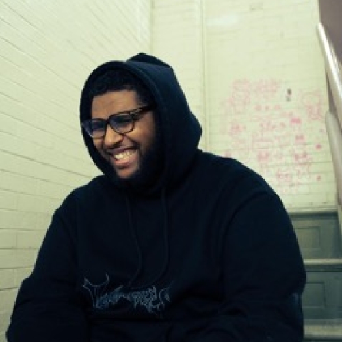 amen ibrahim smiling in a black hoodie and black thick glasses seated in a stairwell. 