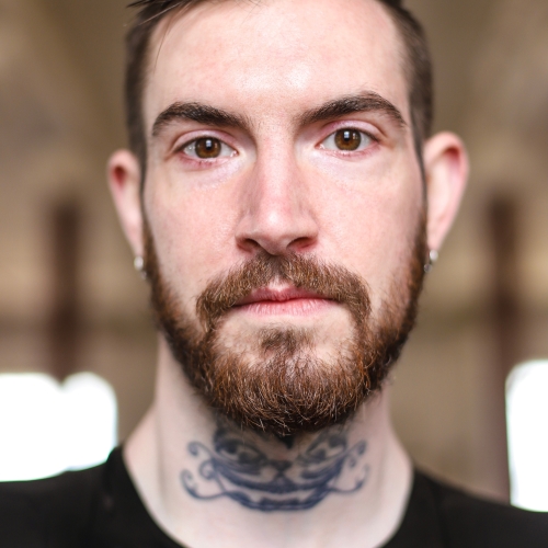 portrait photo of alexander medlin, looking directly into the camera with a neatly trimmed beard, a reflective zippered top, and a wispy tattoo on his neck