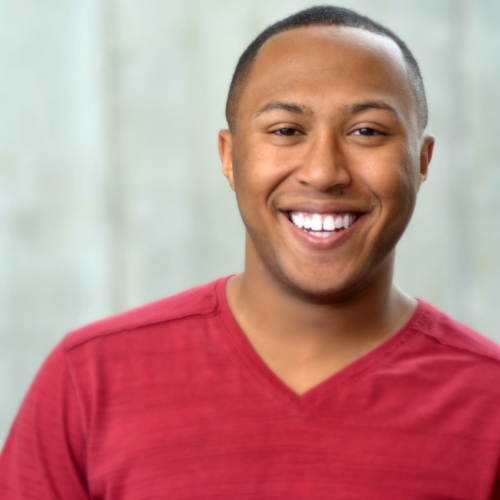 headshot of nathan alford-tate. nathan is wearing a red v neck shirt and is smiling brightly. hathan has short cropped black hair. 
