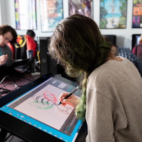 a person with long hair dyed green at the end is seen from their left shoulder in profile working on a digital drawing on a large wide tablet. in the background are additional people working on similar tablets or on computers with their backs turned toward the viewer. 