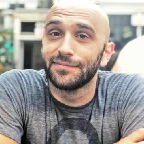 headshot of alex pirani in a grey t shirt with a circle on it. pirani has a scruff beart and is bald and is raising his eyebrows with a small smirk