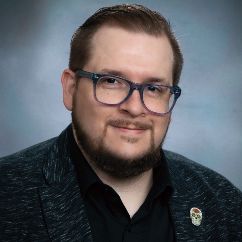 Bradley Philbert, a bearded man in a black shirt and heather grey blazer, smiles and sits for a professional portrait against a marbled grey backdrop. He wears translucent purple glasses and a calavera pin in his lapel.