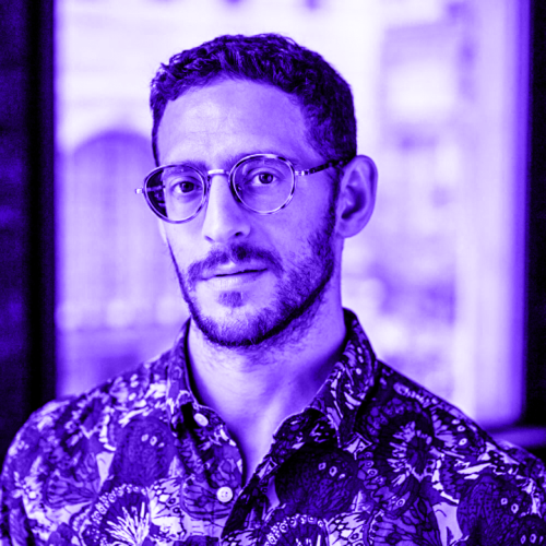 jesse zaritt overlaid with a violet hue. jesse is wearing a densely floral button-up shirt and is looking directly at the viewer from behind tortoise shell round glasses, with a gentle semi-smile from a scruffy face. the background is out of focus. 