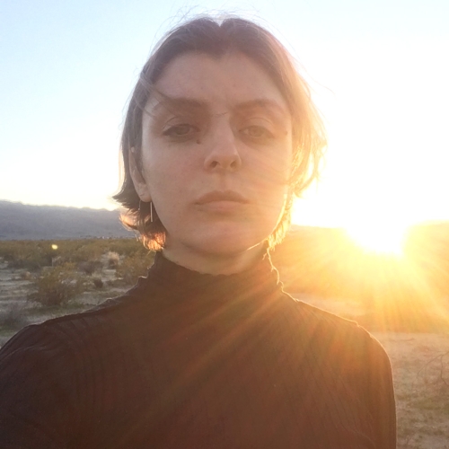 Frank Leasing in a black turtleneck and standing in a desert with the sun shining over their shoulder