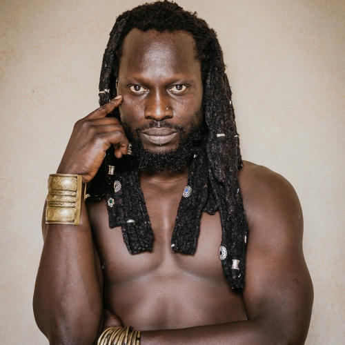 Kaolack stands in front of a light brown background and is shirtless and wearing a large gold cuff on one wrist and a series of gold and black bangles on the other