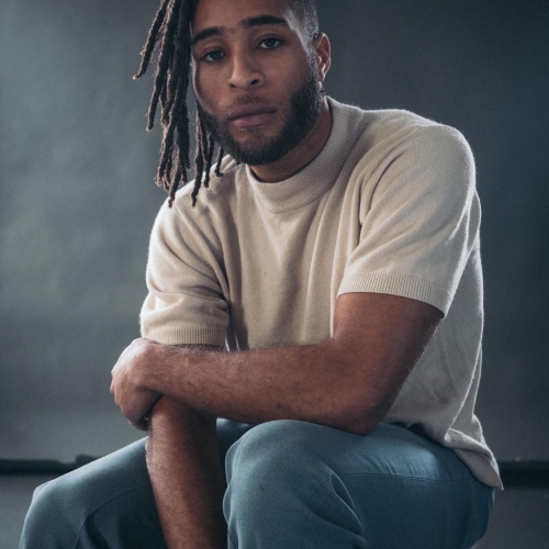 Kingsley Ibeneche sitting in front of a gray and textured backdrop and wearing a beige sweater with short sleeves and jeans