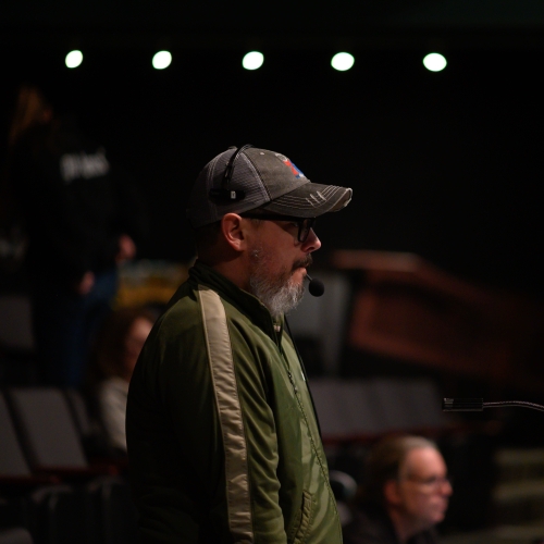 thom weaver seen in profile wearing a green track jacket, gray baseball cap and a headset microphone wrapping around from behind his face to his mouth. He has a gray beard and black glasses, and is in a dimmed theater like space against a background of aisle seating with out of focus people in the background 