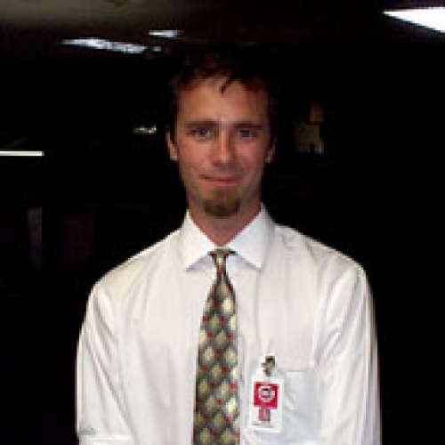 Frank Hartman in white shirt and khaki pants, brown diamond pattern tie, NASA ID badge, holding notebook