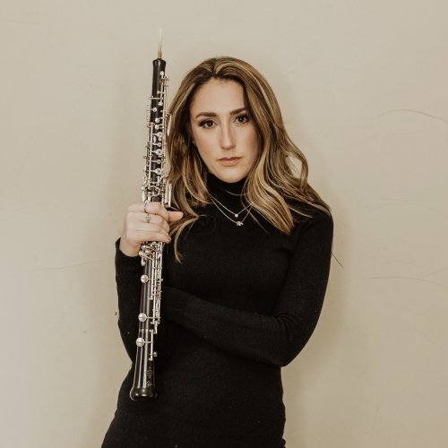 Kaitlyn Walker standing in front of a beige background holding an oboe and dressed in all black