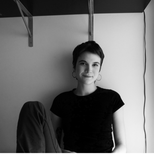 headshot of genevieve wittrock in black and white. genevieve is wearing a black t shirt and large hoop earrings and has a short pixie haircut and is leaning against drywall