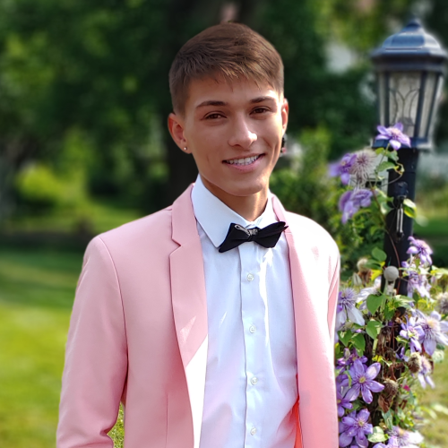 portrait of dennis duran against a lush green background and a tangle of passion flowers on a lamp post. dennis is wearing a pink suit jacket and a black bow tie.