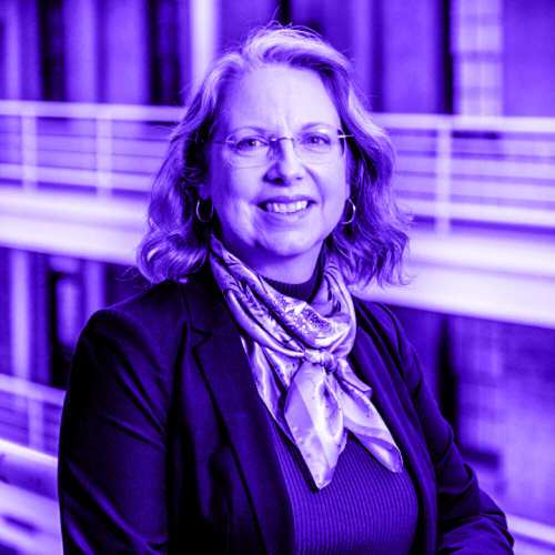 portrait of deborah duffy overlaid with a violet hue. deborah is standing against railing, with railing of the opposing side of the courtyard visible in the back. she is smiling, and is wearing frameless glasses, golden hoop earrings, and a silver scary with a black blazer