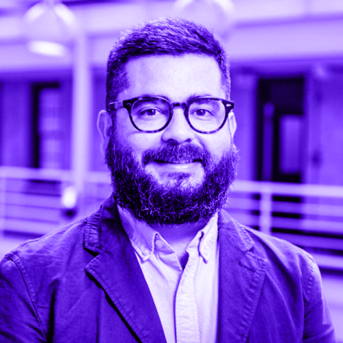 headshot of raul romero overlaid with violet. raul is wearing a blazer and button-up shirt and has a thick full beard and dark horn rimmed tortoise shell glasses with a light smile. 