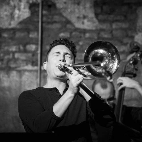 A black and white image of Nick Lombardelli playing trombone in front of a standup bass player with a brick wall behind them