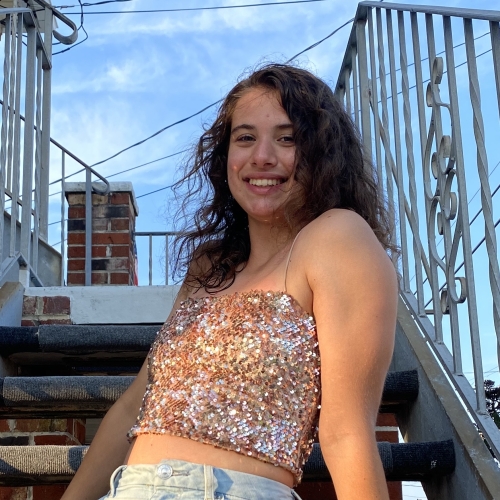 photo of Jamie Seip seen against a background of an outdoor stairwell railing anda blue sky. Jamie is wearing a rose gold sequin top and has long curling brown hair.