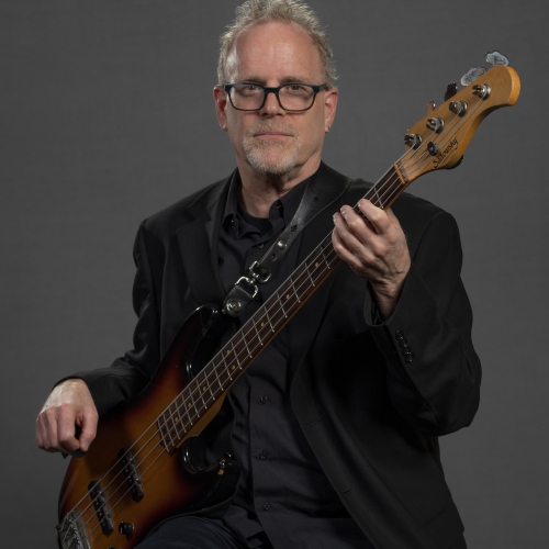 Steve Beskrone in a black suit holding a bass guitar and standing in front of a dark gray background