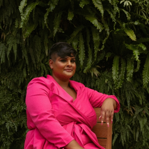 Photo of Preya John, smiling, sitting on a chair in a pink pantsuit with ferns behind her.