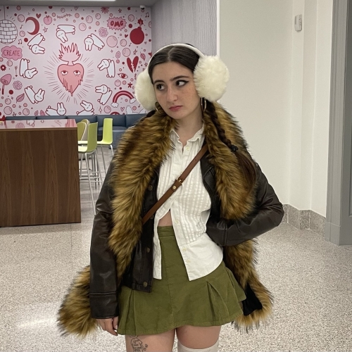 photo of macey baran posing in uarts' student center wearing a short green skirt, and oversize white button up shirt a fur-lined coat and oversized white earmuffs. baran is looking towards the left past a mural depicting cartoonish illustrations in red. 