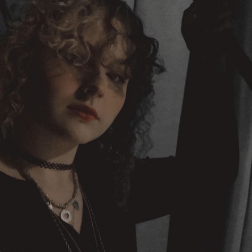 a dimly lit photo of isabel link pictured against a gray background with a black top and delicate chains and curly hair 