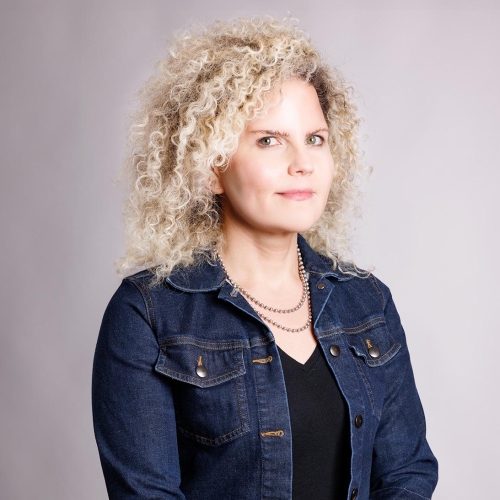 Jennifer Greenburg dressed in a black shirt and a dark blue denim jacket with her hands folded over her lap and posting in front of a light gray and textured backdrop