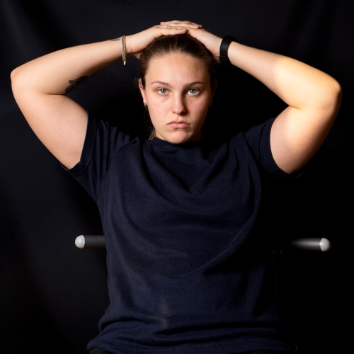 portrait of photographer Jessi Kaufman against a stark black background with her arms raise and her hands behind her head. she is wearing a dark T-shirt.