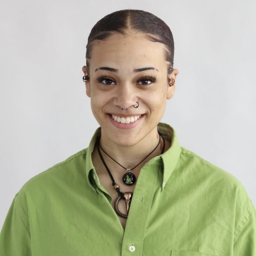 headshot of Katy Robinson. Katy is wearing a green button down shortsleeved shirt with a few pendant poking through at the collar. Katy's hair is pulled back and Katy's eyebrows have notches in the edges. A variety of piercings adorn Katy's ears and nose and a big smile is on Katy's face.